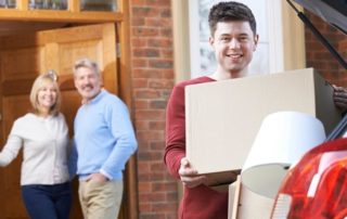 Young man unloads moving boxes from a car, while two smiling adults stand by the front door of a brick house in the background.