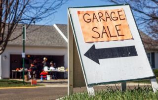 A large Garage Sale sign with a left arrow is placed on a lawn, with a garage sale visible in the background in front of a suburban house.