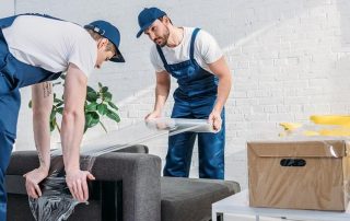 Two movers in blue uniforms wrap plastic around a couch while preparing furniture for moving; a cardboard box is on a nearby white table.