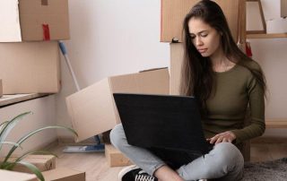 A woman sits on the floor surrounded by cardboard boxes, using a laptop, with a broom and potted plant nearby.