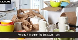 Kitchen items, including mugs, bowls, and a teapot, are being packed with brown paper and placed into cardboard boxes on a countertop.